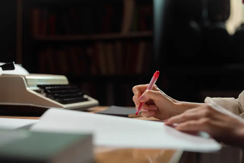 Author marking up a manuscript in red pen beside a typewriter